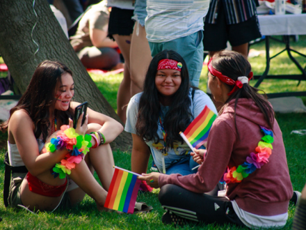 Group of women sitting on grass wearing rainbow apparel and holding rainbow flags during Pride on the Plaza in Concord CA