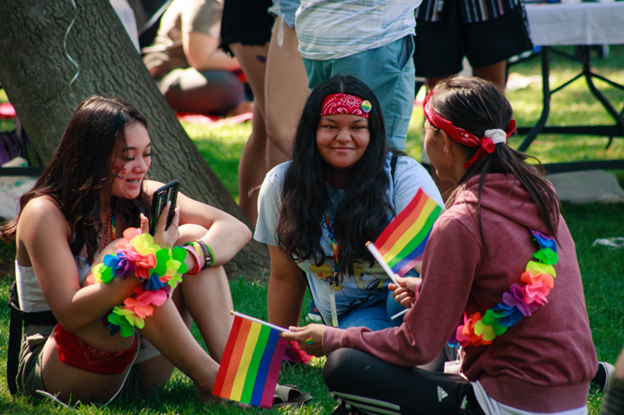 Group of women sitting on grass wearing rainbow apparel and holding rainbow flags during Pride on the Plaza in Concord CA
