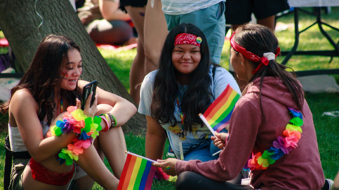 Group of women sitting on grass wearing rainbow apparel and holding rainbow flags during Pride on the Plaza in Concord CA