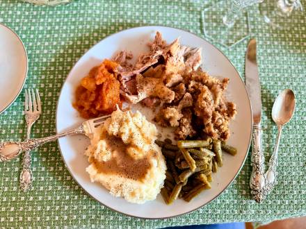 A white plate on a green tablecloth is filled with turkey, green beans, mashed potatoes, and more Thanksgiving goodies.