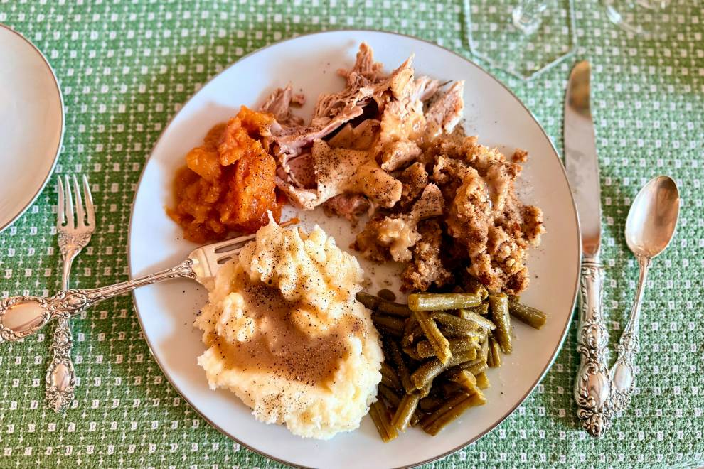 A white plate on a green tablecloth is filled with turkey, green beans, mashed potatoes, and more Thanksgiving goodies. 